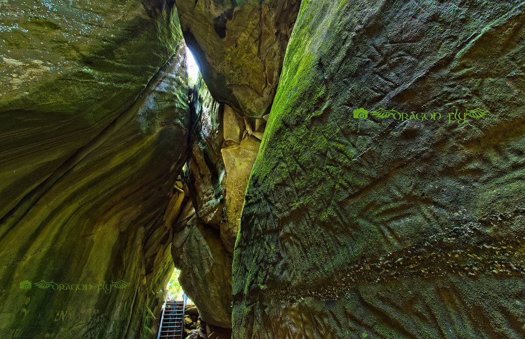 Ancient rock carved with prehistoric symbols and human figures inside a dramatic stone fissure, dappled light from above